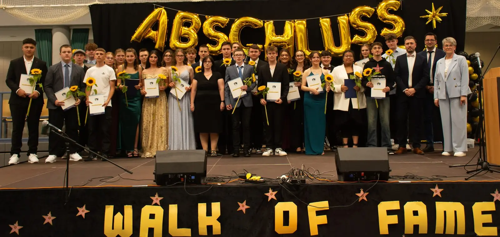 Große Abschlussgruppe mit Zeugnissen und Sonnenblumen auf der Bühne über dem Banner „WALK OF FAME“