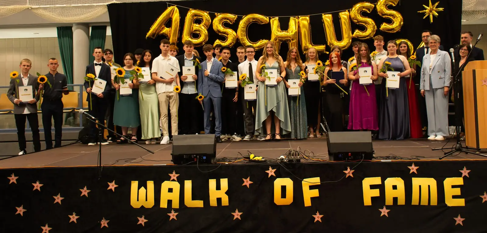 Weitere große Abschlussgruppe mit Zeugnissen auf der „WALK OF FAME“-Bühne, rechts ein Rednerpult