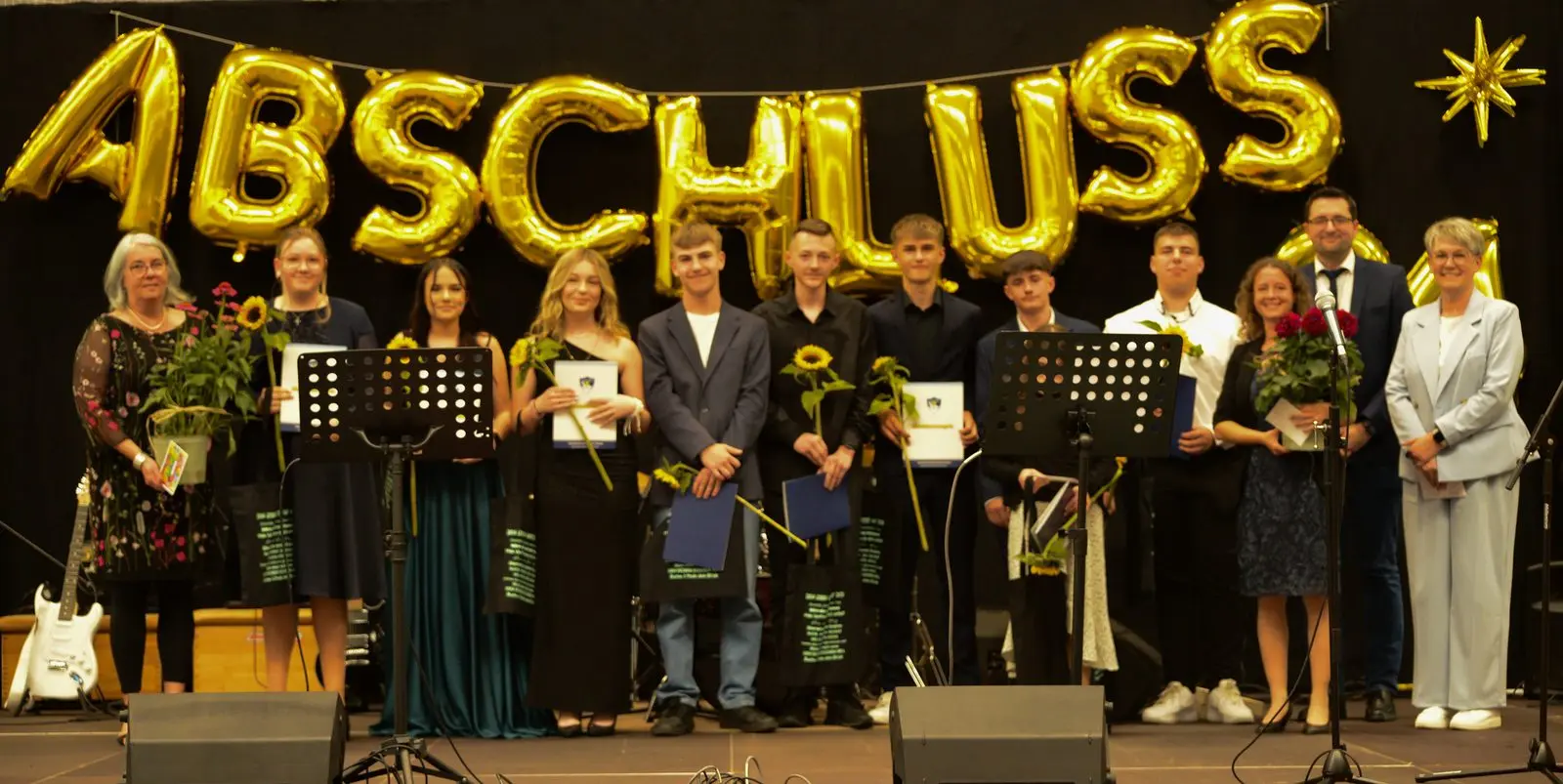 Abschlussgruppe mit Sonnenblumen und Mappen auf der Bühne unter goldenen Ballonbuchstaben „ABSCHLUSS“, außen Erwachsene mit Blumen