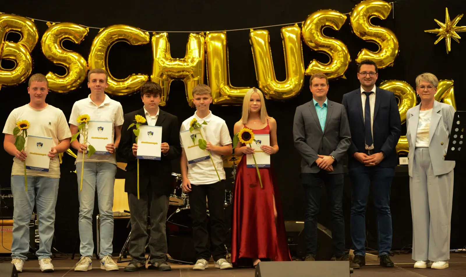 Fünf Abschlussschülerinnen und -schüler mit Sonnenblumen und Zeugnismappen, daneben drei Erwachsene auf der Bühne