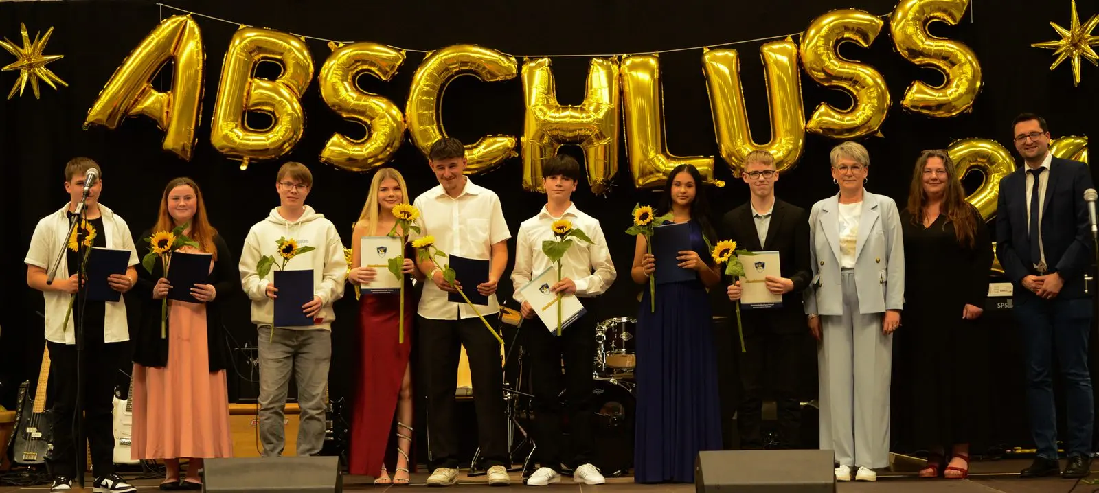 Acht Abschlussschülerinnen und -schüler mit Sonnenblumen und Mappen, links einer am Mikrofon, rechts drei Erwachsene