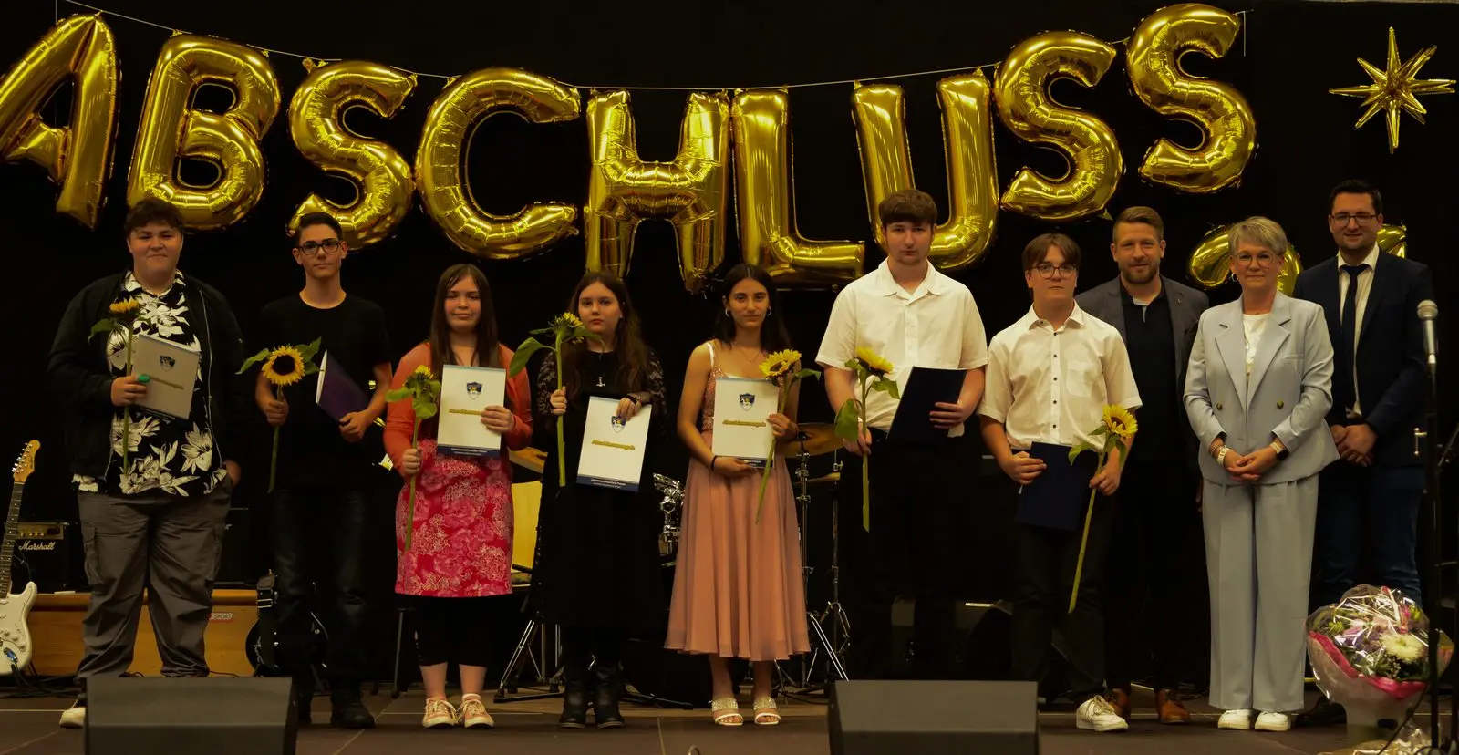 Acht Abschlussschülerinnen und -schüler mit Sonnenblumen und Zeugnissen neben drei Erwachsenen, vorn rechts ein Blumenstrauß