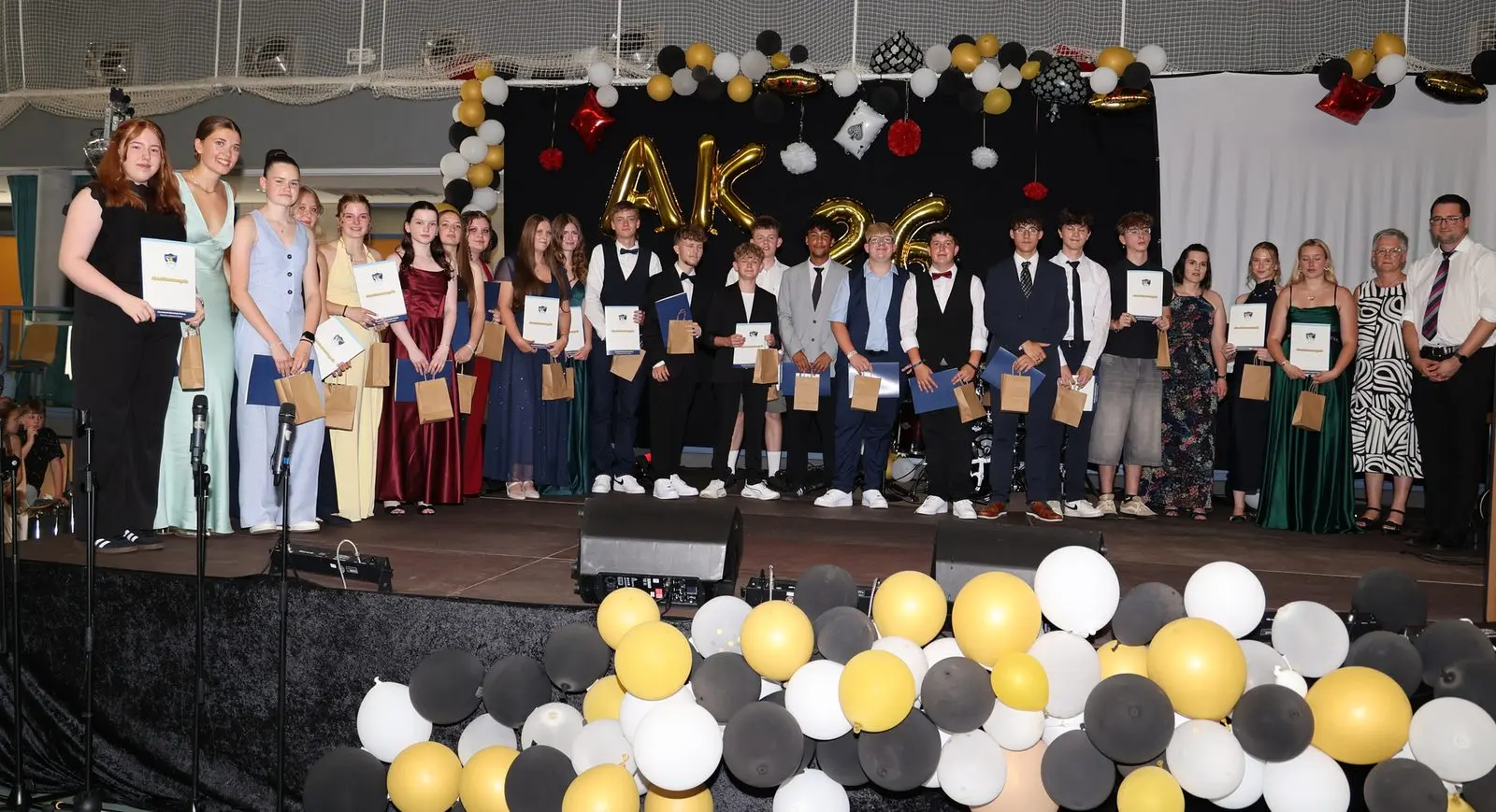 Große Abschlussgruppe mit Zeugnissen und Geschenktüten auf der Bühne, vorn schwarz-weiß-gelbe Ballons