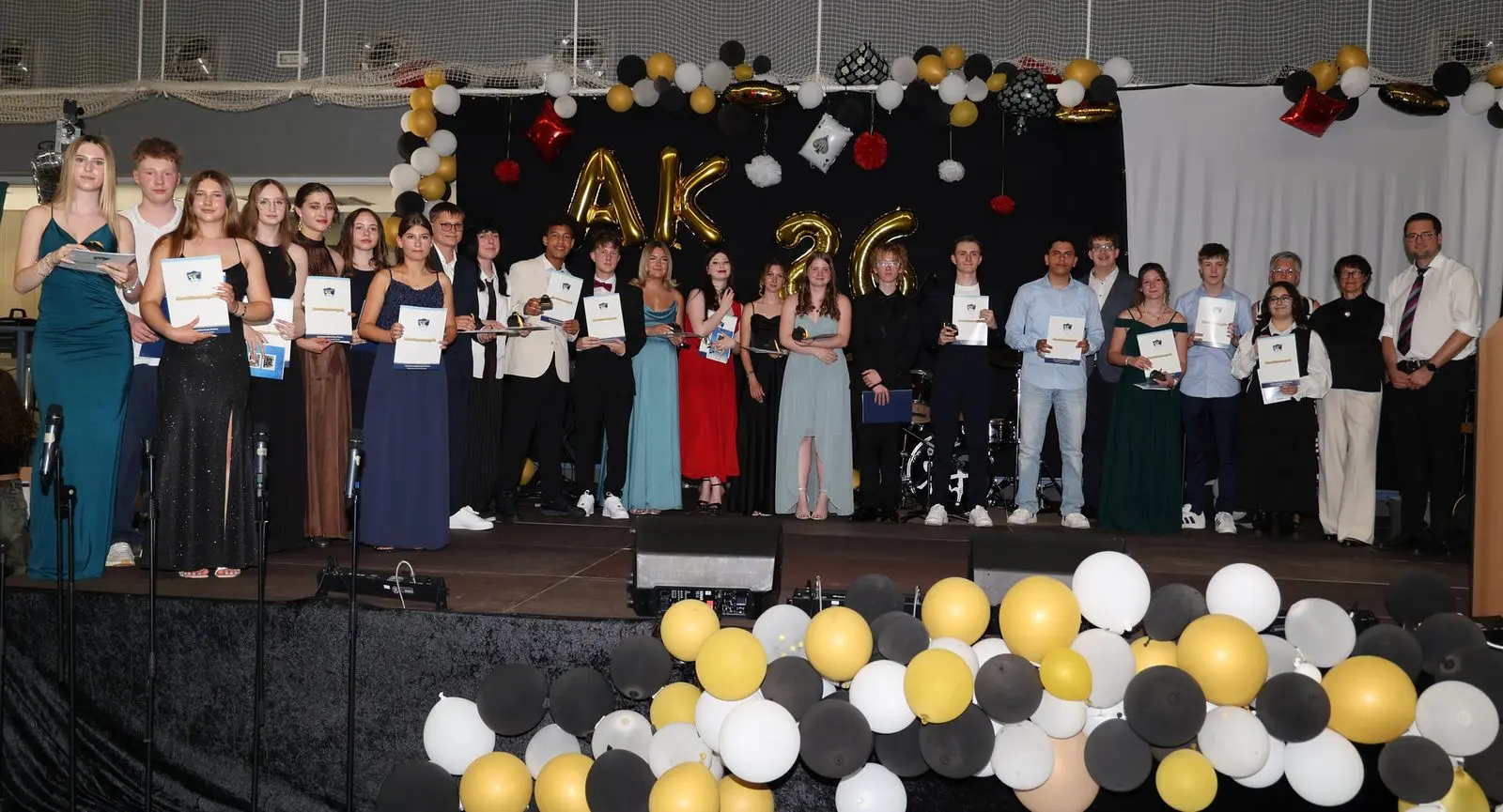 Große Abschlussgruppe mit Zeugnissen auf der Bühne, links eine Schülerin im türkisfarbenen Kleid