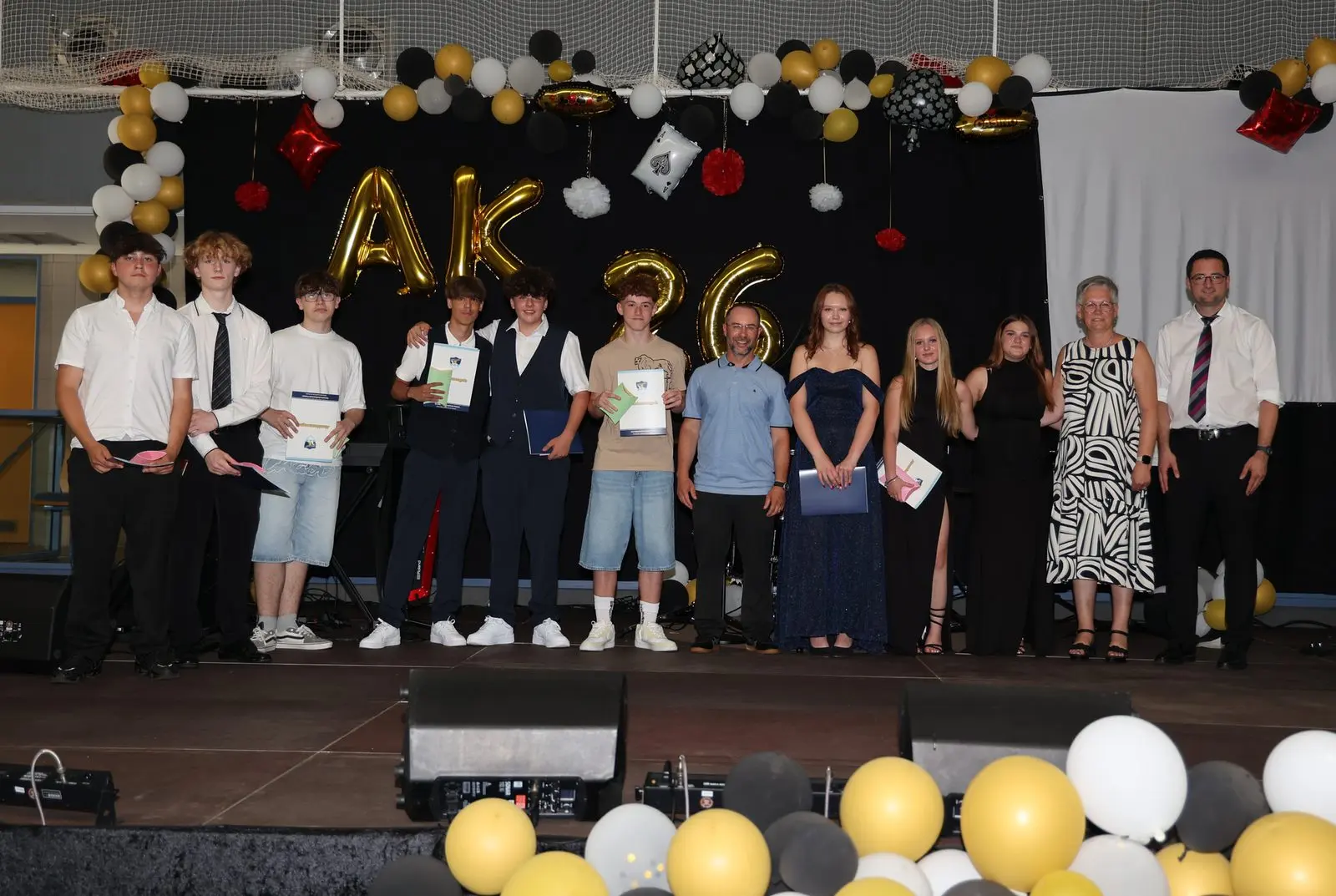 Abschlussgruppe mit Zeugnissen auf der Bühne vor „AK 26“-Ballons, rechts drei Erwachsene