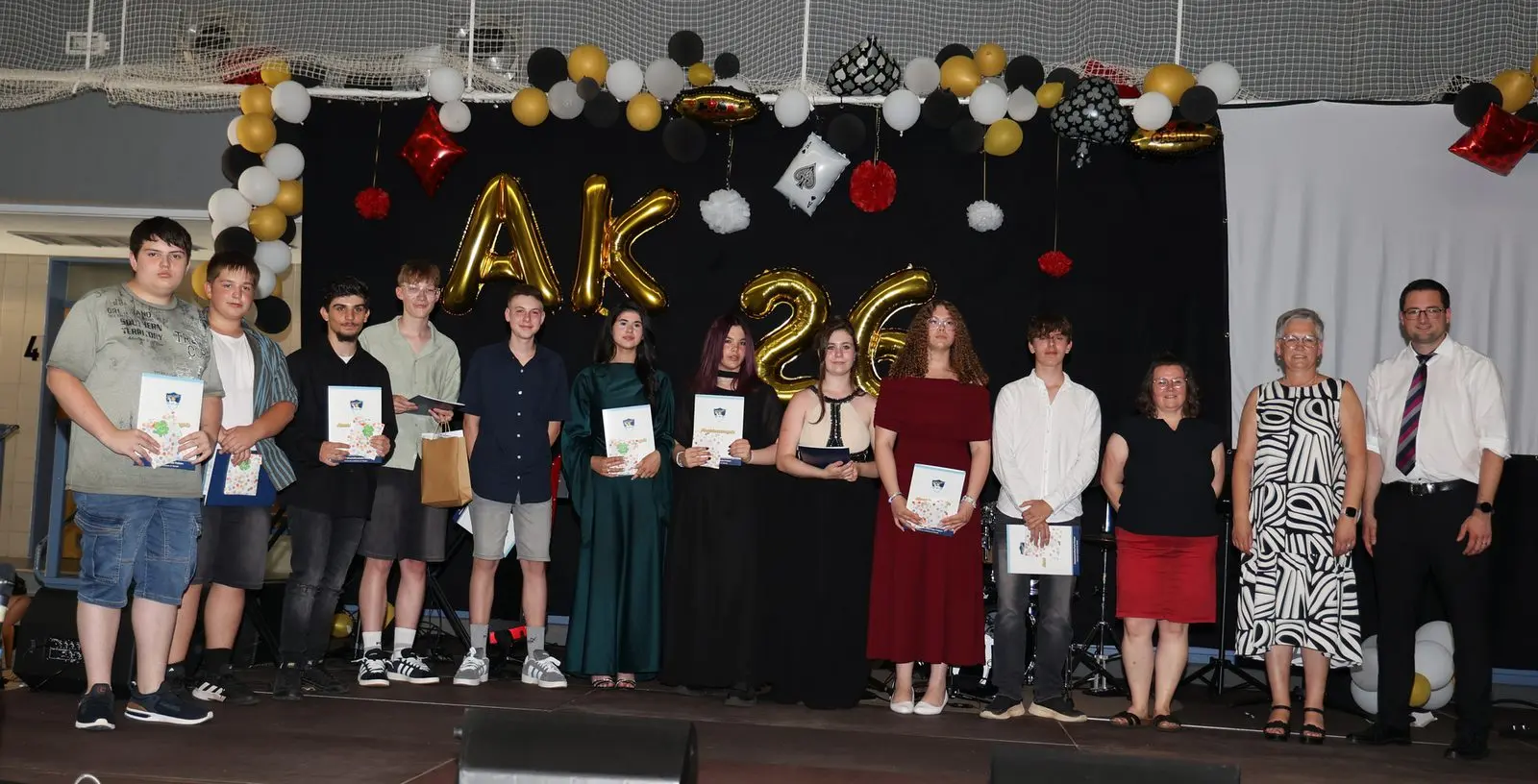 Elf Abschlussschülerinnen und -schüler mit Zeugnissen und drei Erwachsene vor dem „AK 26“-Schriftzug
