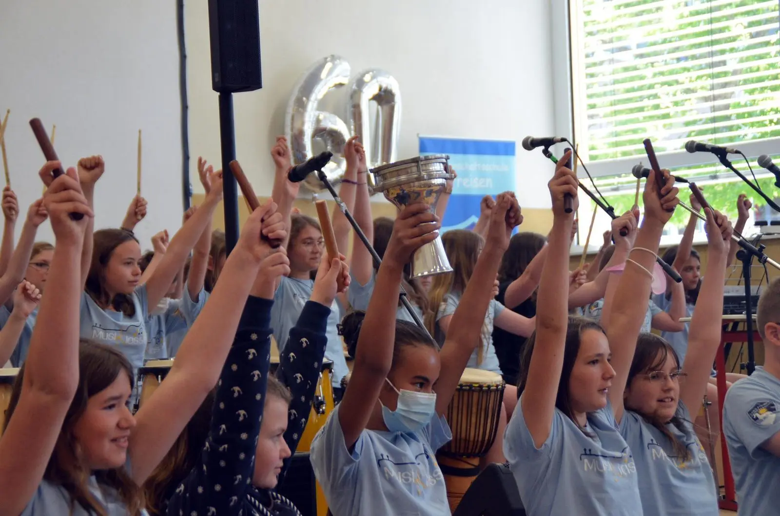 Schülerinnen der Musikklasse heben jubelnd ihre Trommelstöcke, im Hintergrund Ballons in Form der Zahl 60