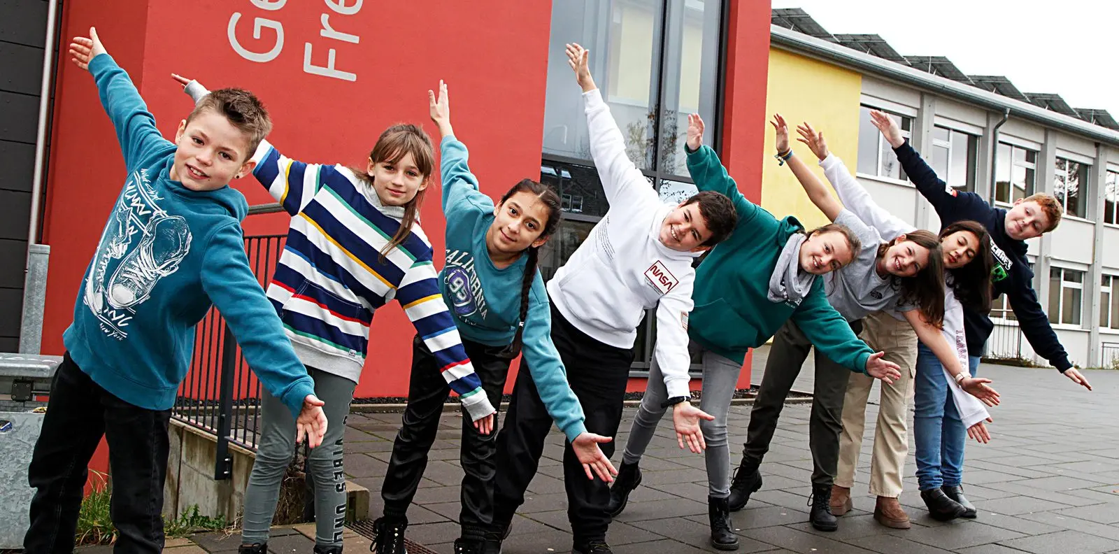 Schülerinnen und Schüler stehen in einer Reihe vor dem Schulgebäude und lehnen sich gemeinsam zur Seite