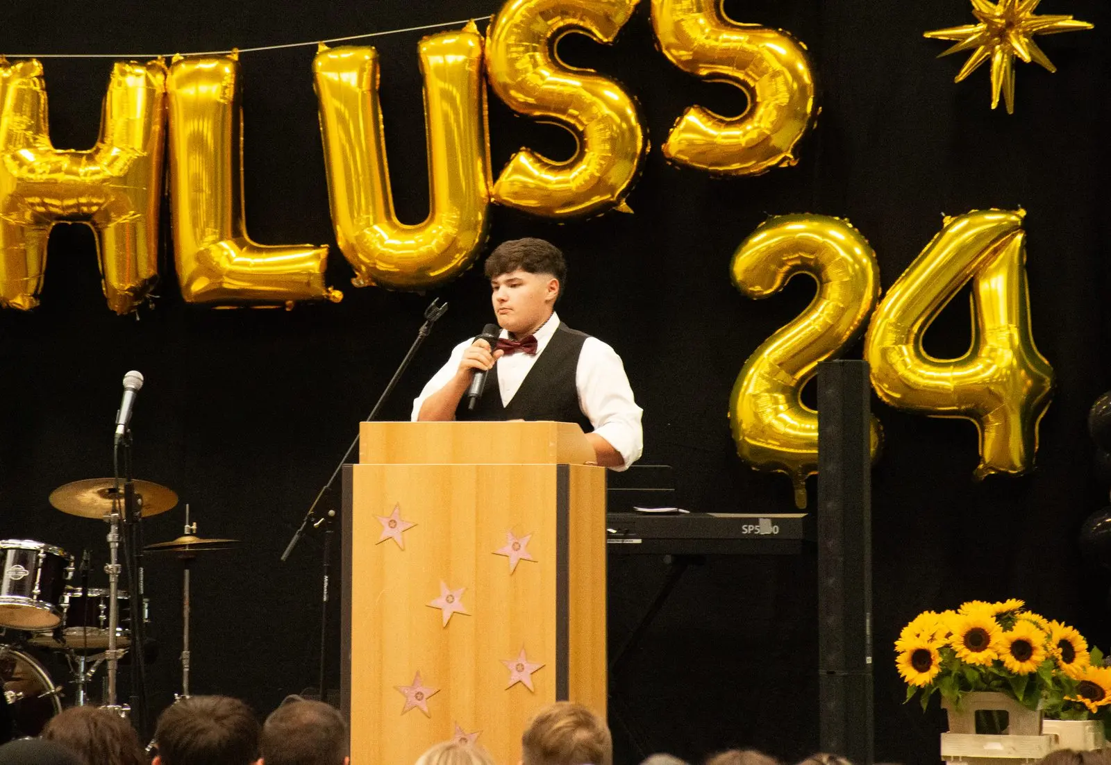 Ein Schüler mit Weste und Fliege moderiert am Rednerpult, rechts Sonnenblumen