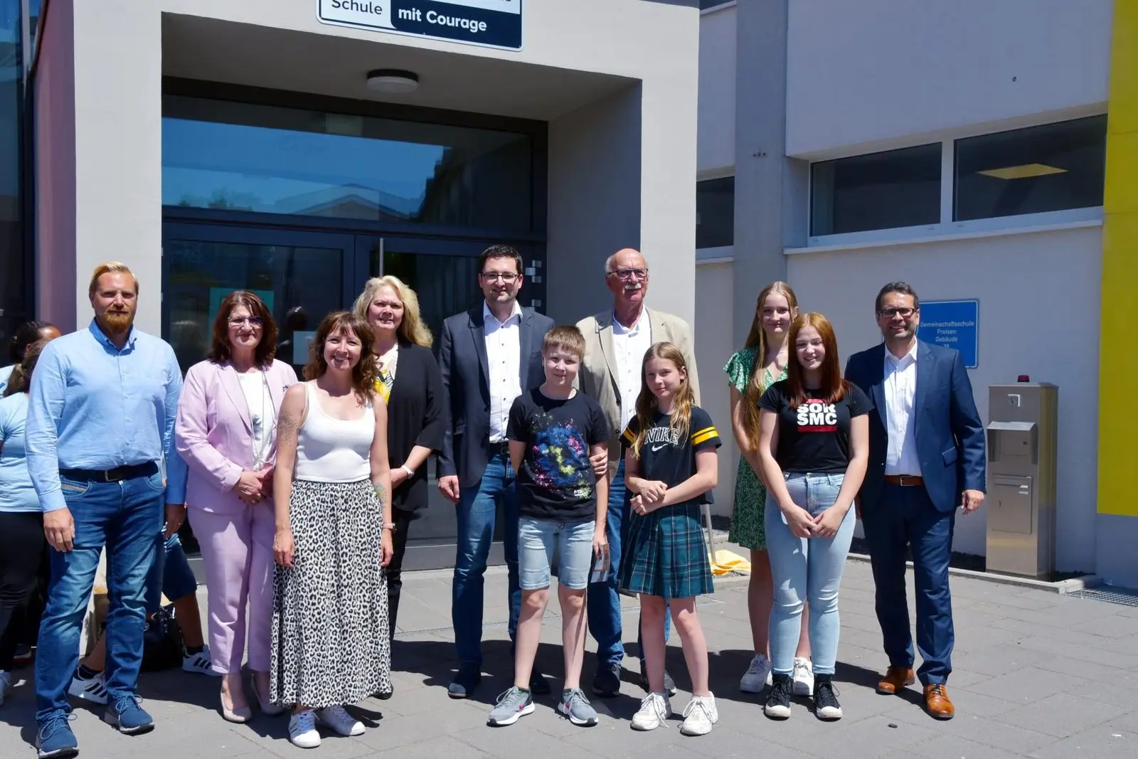Gruppenfoto von Erwachsenen sowie Schülerinnen und Schülern vor dem Schuleingang mit dem Schild „Schule ohne Rassismus – Schule mit Courage“