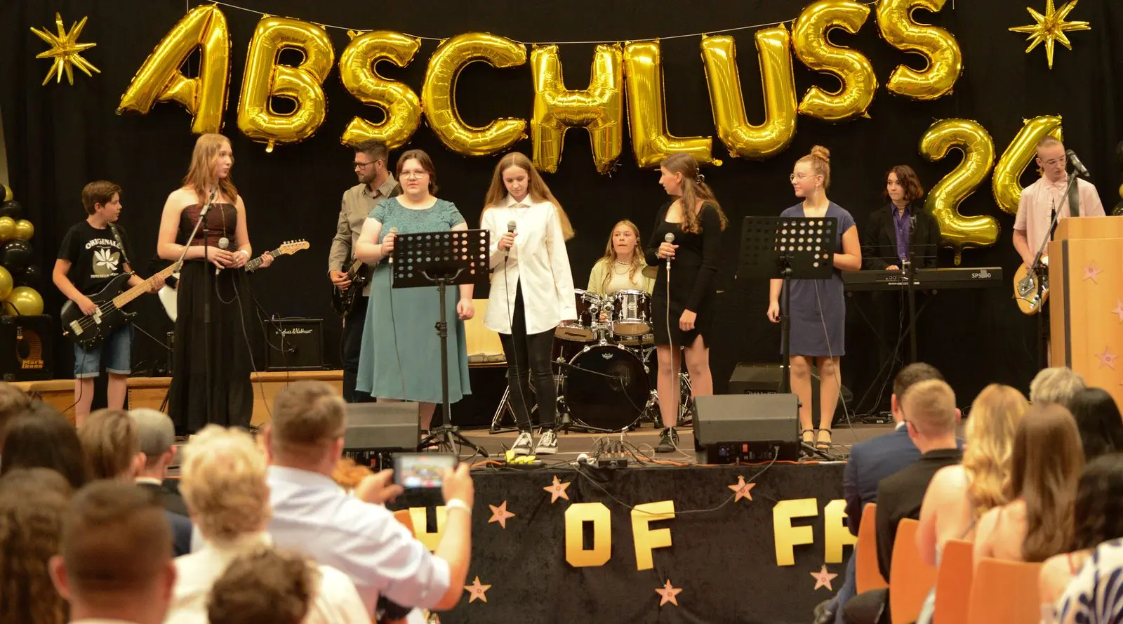 Schulband mit mehreren Sängerinnen auf der Bühne, im Vordergrund Publikum, eine Person filmt mit dem Handy