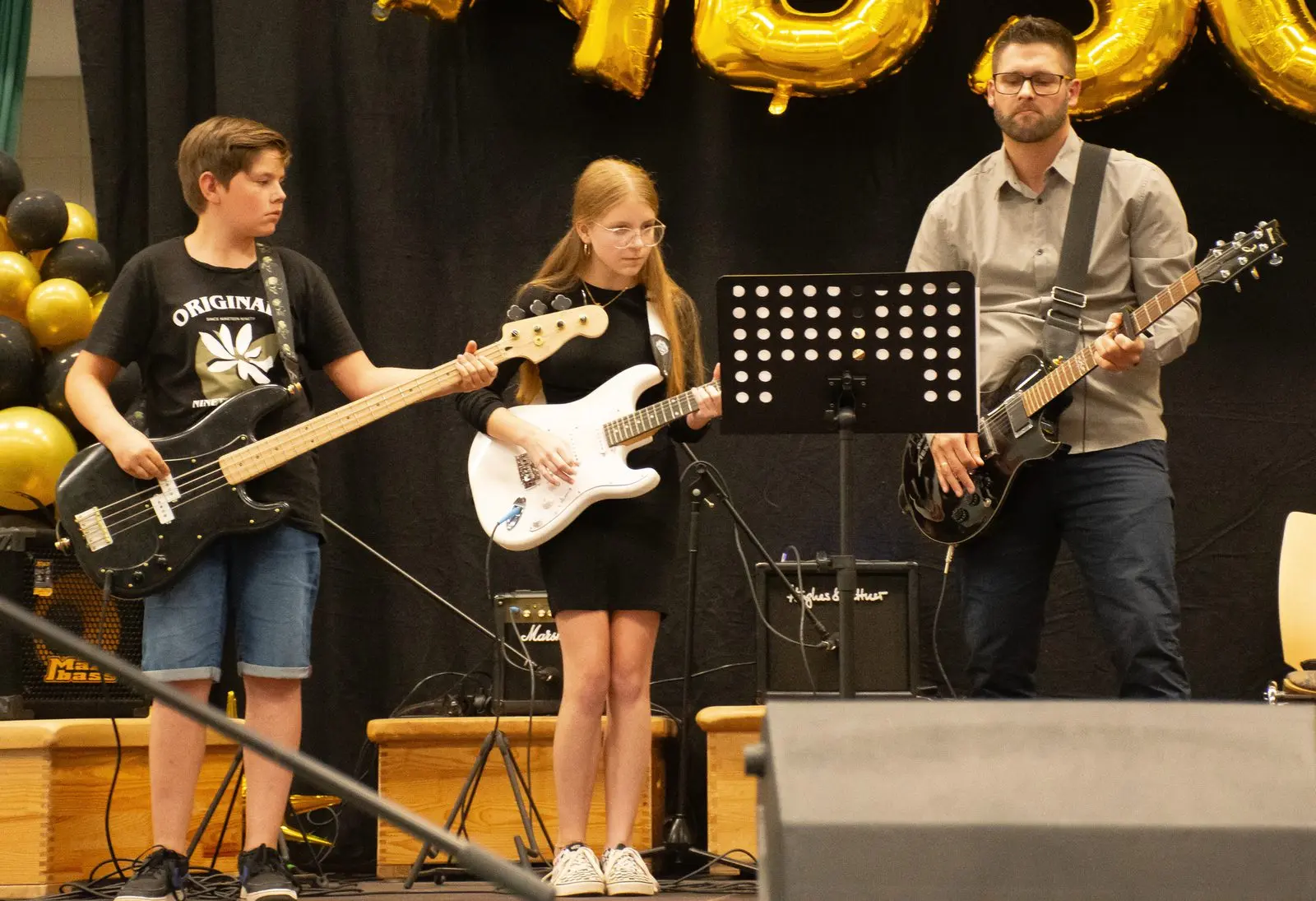 Drei Gitarristen der Schulband: ein Schüler am Bass, eine Schülerin mit weißer E-Gitarre und ein Erwachsener