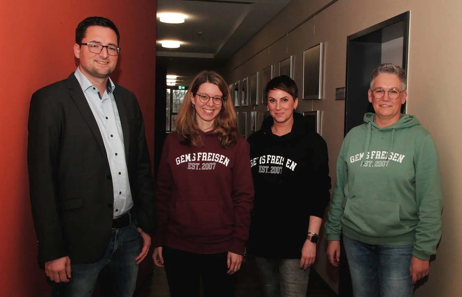Vier Personen im Schulflur, drei tragen Pullover mit der Aufschrift „GEMS FREISEN EST. 2007“