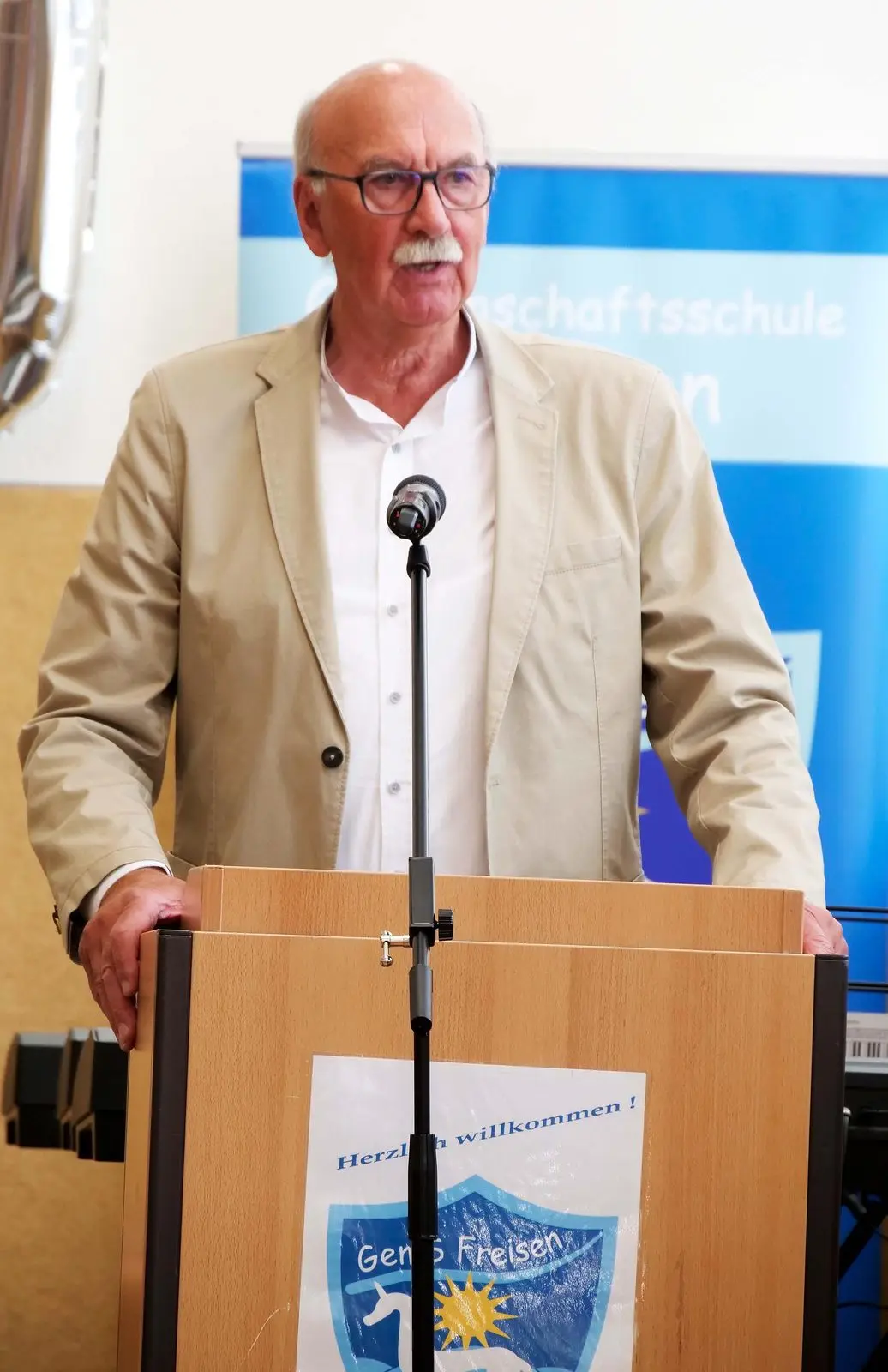 Älterer Mann spricht am Rednerpult mit dem Schullogo