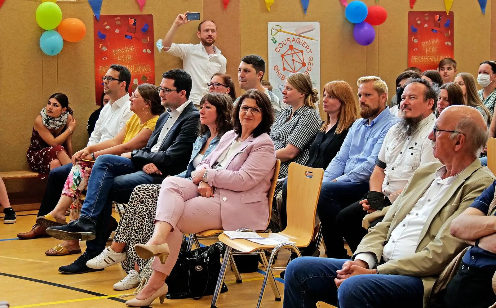 Gäste in Stuhlreihen vor bunt geschmückter Wand