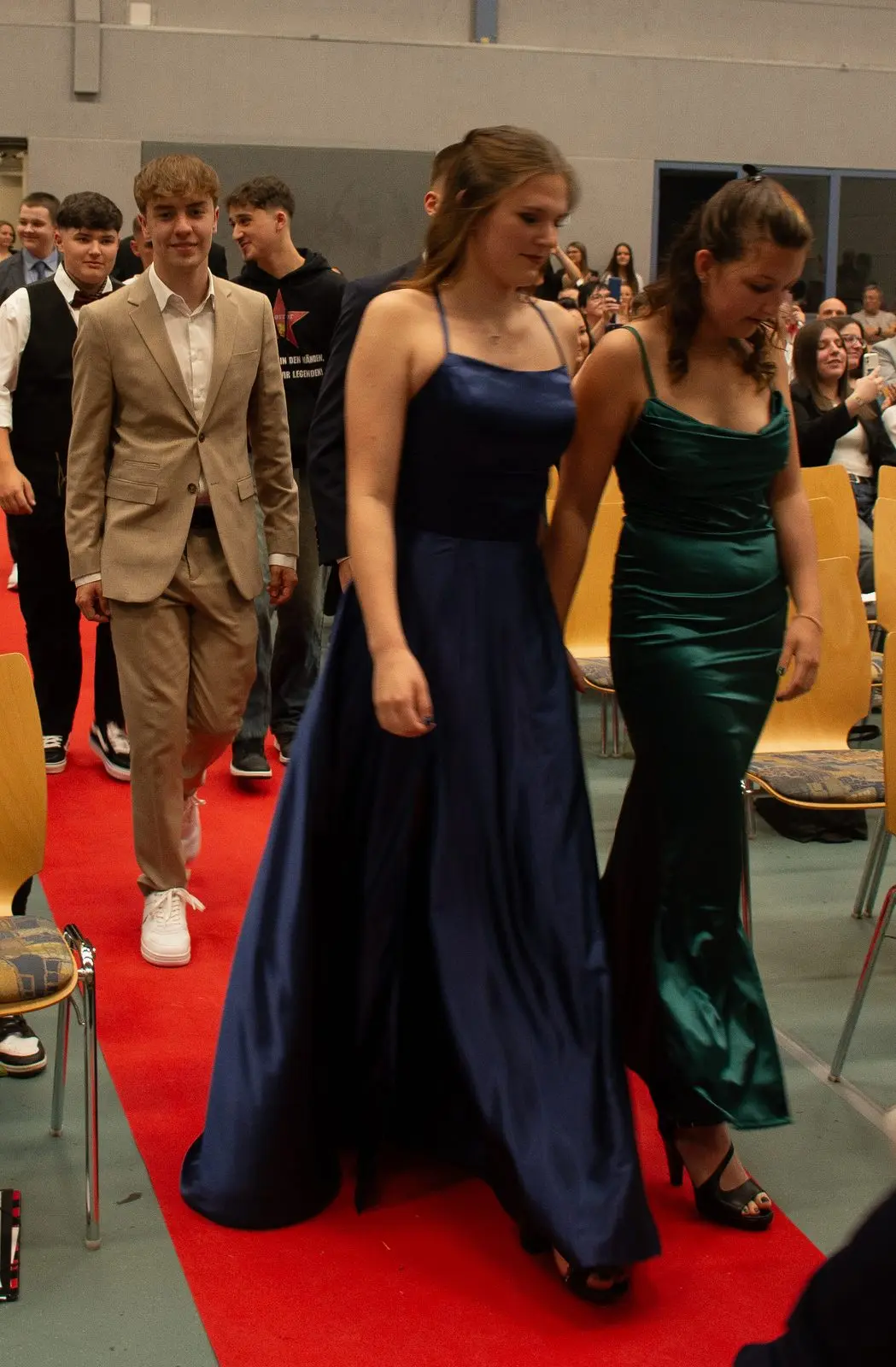 Abschlussschülerinnen und -schüler in festlicher Kleidung ziehen paarweise über einen roten Teppich in die Halle ein