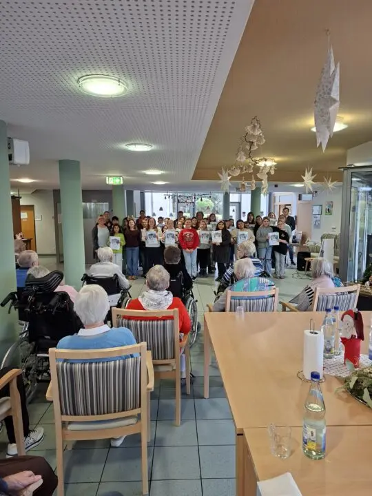 Schülergruppe singt mit Liedblättern im Aufenthaltsraum vor Seniorinnen und Senioren