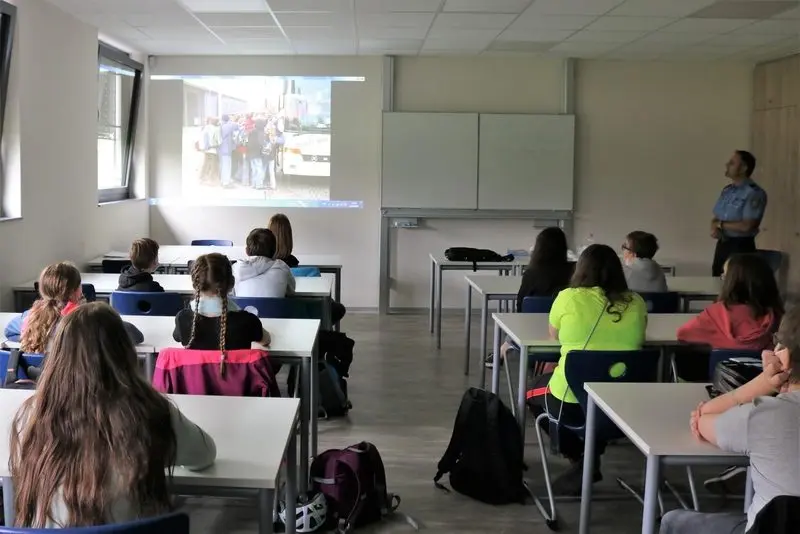 Schülerinnen und Schüler im Klassenraum schauen auf eine Projektion zur Busschule, vorne ein Polizist