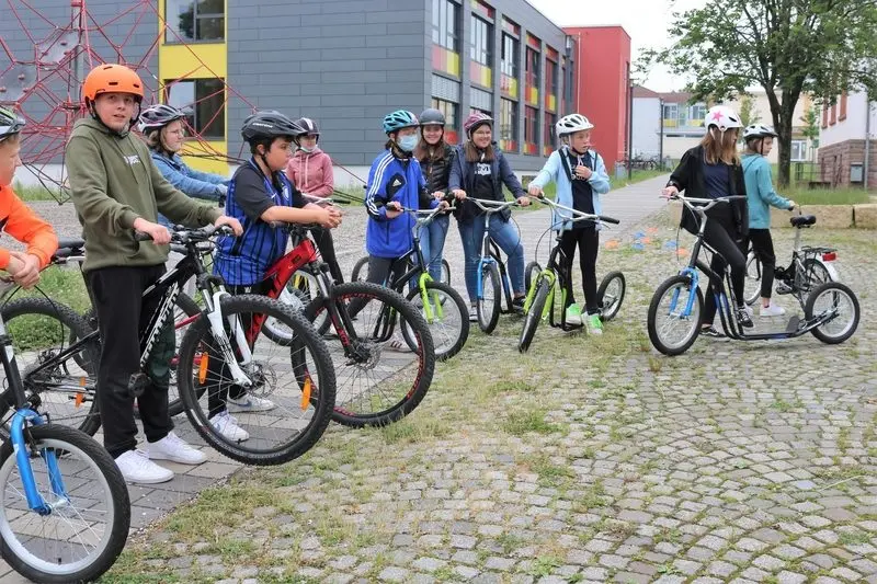 Schülerinnen und Schüler mit Helmen stehen mit Fahrrädern und Tretrollern auf dem Schulhof