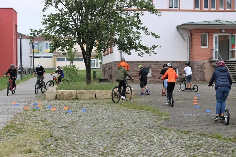 Schülerinnen und Schüler mit Helm fahren auf dem Schulhof Fahrrad und Tretroller durch einen Hütchenparcours