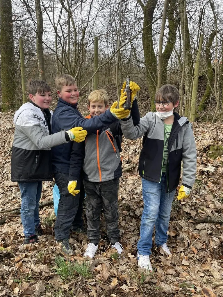 Vier Jungen mit Arbeitshandschuhen halten im Laubwald eine gefundene Flasche hoch