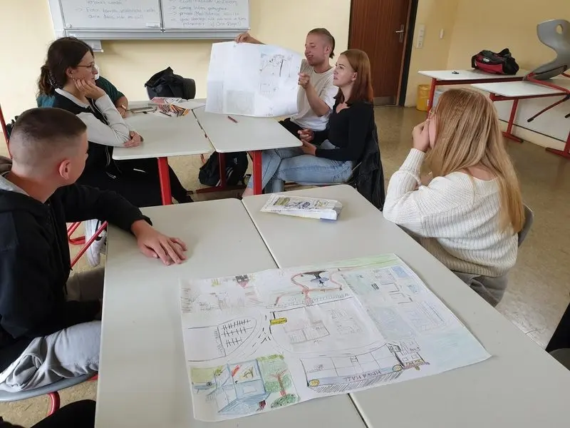 Jugendliche am Gruppentisch mit selbst gezeichneten Stadtplänen, einer hält einen Plan hoch