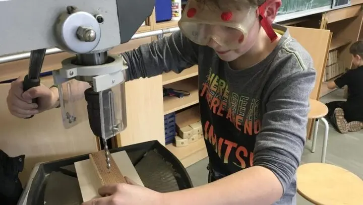 Schüler mit Schutzbrille bohrt an einer Ständerbohrmaschine ein Loch in ein Holzstück
