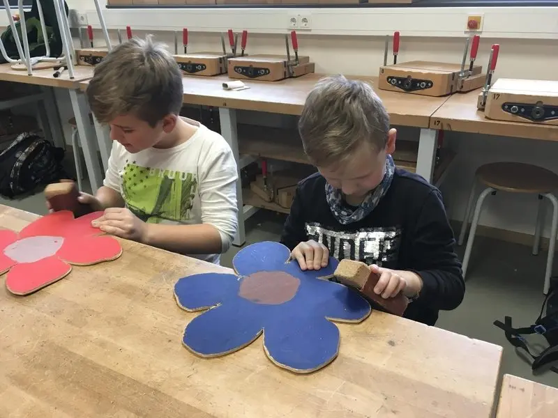 Zwei Schüler schleifen rot und blau bemalte Holzblumen an einer Werkbank