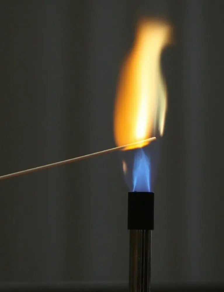 Holzstab in einer Bunsenbrennerflamme, die blaue Flamme färbt sich oben orange