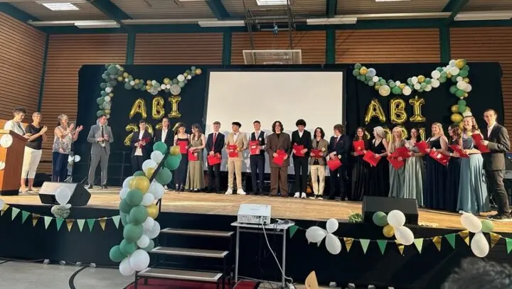 Abiturientinnen und Abiturienten mit roten Zeugnismappen auf der Bühne, Erwachsene applaudieren