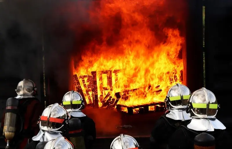 Feuerwehrleute mit silbernen Helmen vor einem lodernden Brand in einer Brandübungsanlage