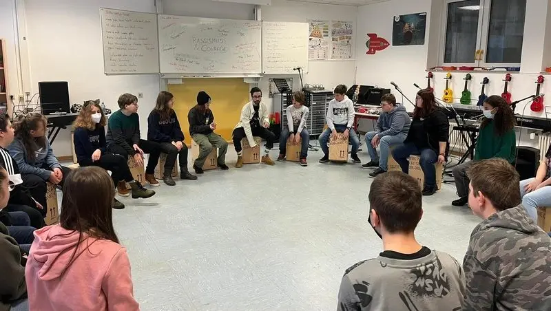 Jugendliche sitzen im Musikraum im Kreis auf Cajóns, an der Tafel steht „Rassismus & Courage“