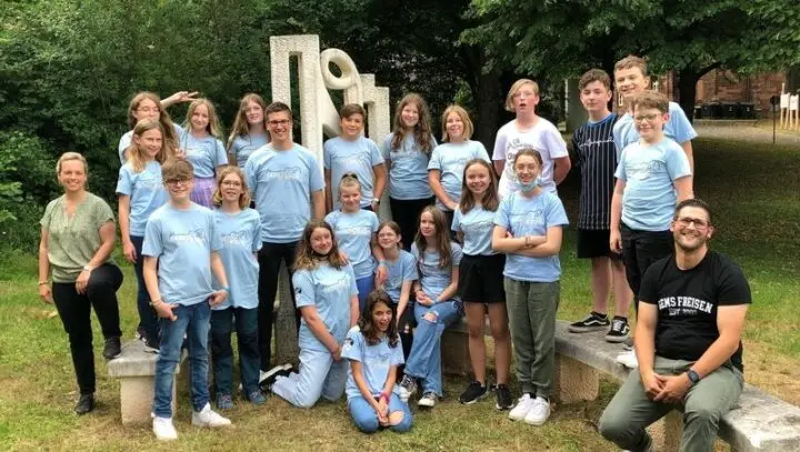Gruppenfoto der Klasse in hellblauen T-Shirts mit zwei Erwachsenen vor einer Skulptur im Grünen