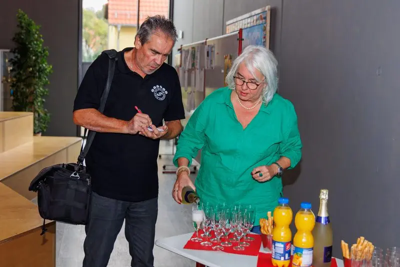 Ein Mann notiert etwas, daneben schenkt eine Frau an einem Tisch mit Gläsern und Saftflaschen Sekt ein