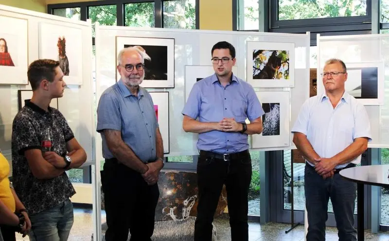 Ein Redner spricht vor Stellwänden mit gerahmten Fotos, daneben drei Zuhörer