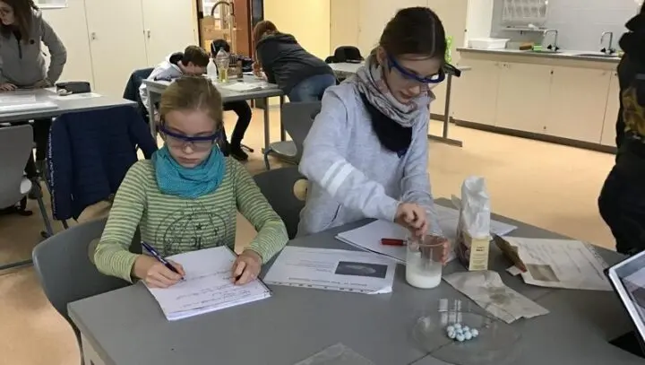 Zwei Schülerinnen mit Schutzbrillen beim Experimentieren und Protokollieren im Fachraum