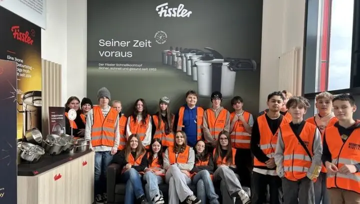 Schülergruppe in orangefarbenen Besucherwesten im Fissler-Showroom vor einer Wand mit Schnellkochtöpfen