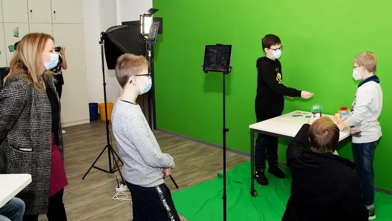 Schüler filmen im Makerspace vor einem Greenscreen mit Tablet und Studioleuchte, Erwachsene schauen zu