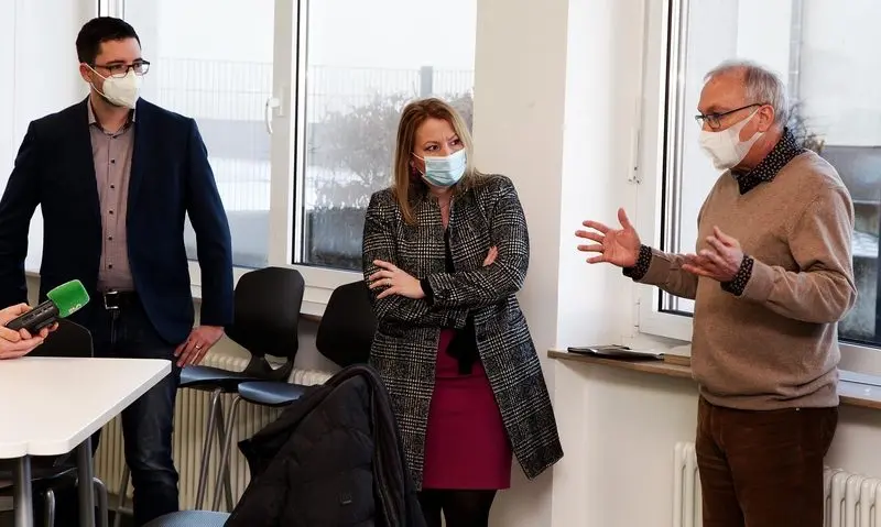 Drei Erwachsene mit Masken im Gespräch, ein Mann erklärt gestikulierend, ein Mikrofon ist auf ihn gerichtet