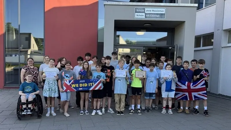 Große Gruppe mit Urkunden, Pokalen, britischer Flagge und Banner „We did it!“ vor dem Schuleingang