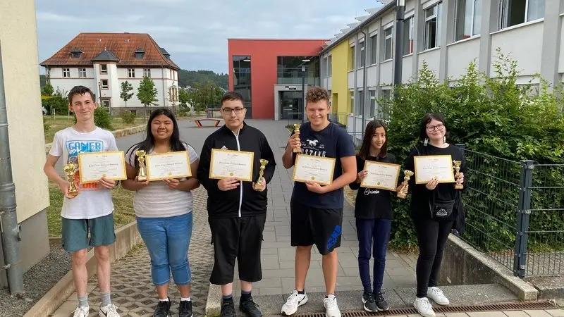 Sechs Schülerinnen und Schüler mit Urkunden und Pokalen vor dem Schulgebäude