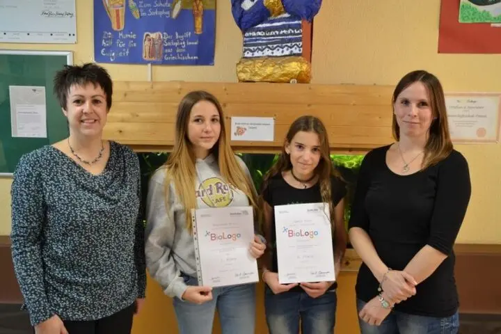 Zwei Schülerinnen mit BioLogo-Urkunden zwischen zwei Frauen vor einem Aquarium
