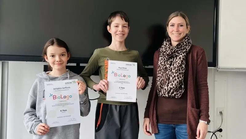 Zwei Kinder zeigen ihre BioLogo-Urkunden für den 8. und 3. Platz, daneben eine Erwachsene
