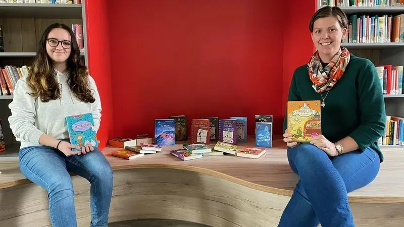 Zwei Personen halten in der Bibliothek Bücher hoch, zwischen ihnen die gespendeten Bücher