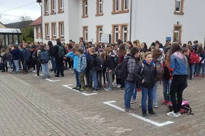 Viele Schülerinnen und Schüler mit Rucksäcken warten an markierten Aufstellflächen vor einem Schulgebäude