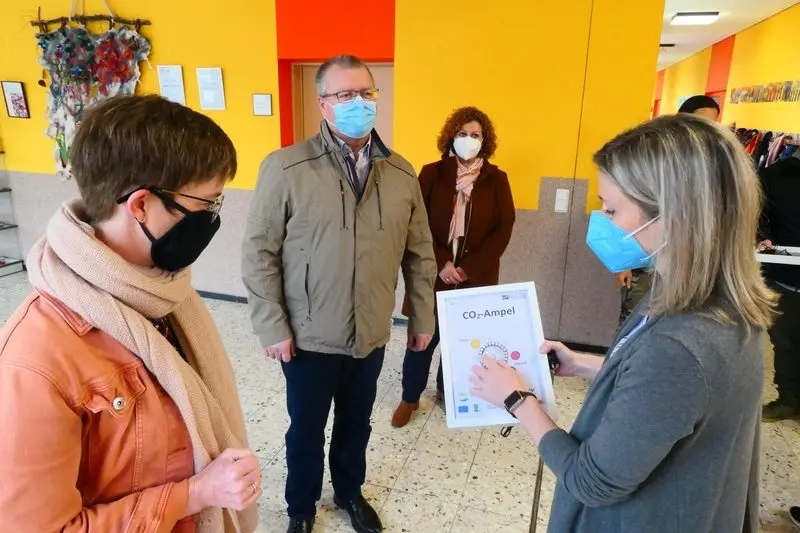 Frau mit Maske zeigt eine gerahmte CO2-Ampel, drei Erwachsene schauen zu