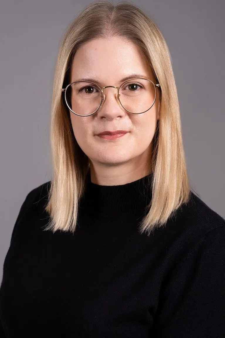 Porträt einer Frau mit blonden Haaren und Brille vor grauem Hintergrund