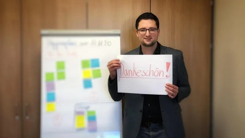 Ein Mann hält ein Blatt mit der Aufschrift „Dankeschön!“ hoch, daneben ein Flipchart