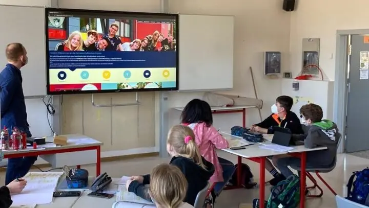 Lehrer und Kinder mit Masken im Klassenraum vor einer digitalen Tafel mit der Schulwebsite