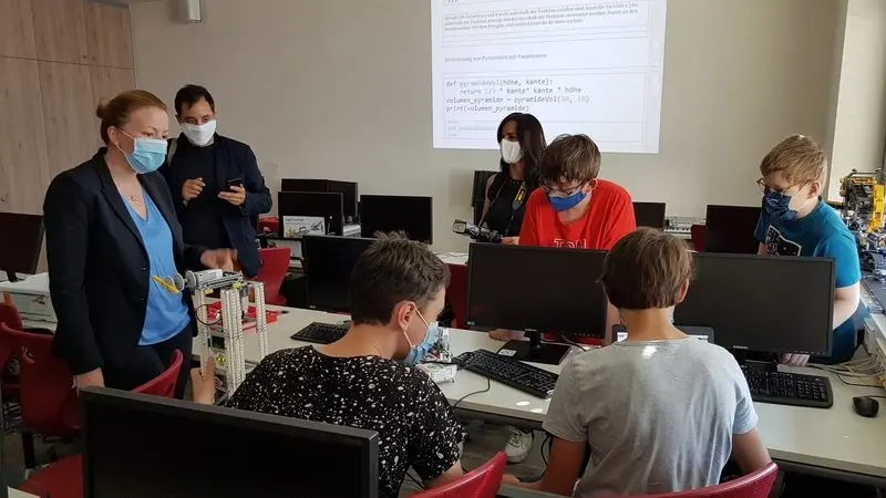 Besucherin spricht mit Jugendlichen an Computern, an der Wand Python-Code